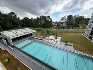 Aerial view of an outdoor pool area with turquoise water, white lounge chairs, a small playground slide, and surrounding trees and buildings.