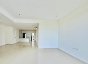 Bright, empty open-plan living area with white walls and glossy tile floor, leading to a modern kitchen with a breakfast bar in the back.