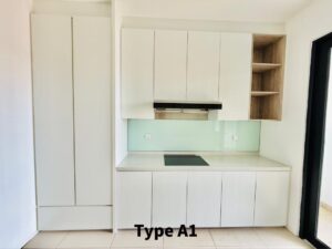 Compact white kitchen with a tall built-in cabinet on the left, white upper and lower cabinets, and a range hood above a cooktop. A light green glass backsplash runs along the wall, with open wooden shelves on the right side.