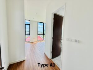 Bright empty living area with light wood floors, white walls, and tall windows showing a city view; a dark wood door is ajar to the right.