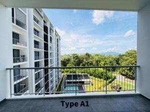 Balcony view of a white multi-story apartment building with a railing, overlooking a pool, green lawn, and distant hills under a blue sky.