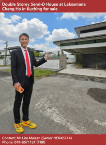 Real estate agent in a dark suit and red tie points toward a modern house for sale, holding a phone, on a sunny day.