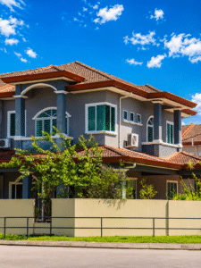 Two-story gray house with a red-tiled roof, green-framed windows, and a beige boundary wall on a sunny day, with blue sky.