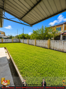Sunny backyard with a green lawn, a concrete patio edge, and a white fence with yellow posts; houses and trees beyond.