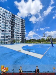 Wide shot of a modern multi-story apartment building on the left, bright blue sky with clouds, and a blue outdoor basketball court in the foreground with a hoop on the right; banners at the bottom.