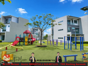 Playground in a sunny residential complex with a red slide, swings, and a central tree surrounded by modern gray buildings.