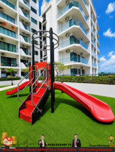 Red plastic slide with a climbing stairs structure on bright green artificial turf, set against white high-rise apartment buildings on a sunny day.