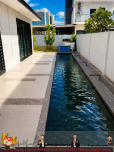 Narrow rectangular pool along a white-walled patio, with beige tiled deck on the left and modern buildings in the background under a blue sky.