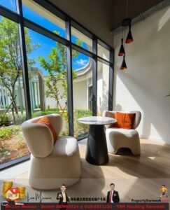 Bright lounge area with two beige rounded armchairs, orange cushions, and a marble-topped round table beside large windows showing greenery.