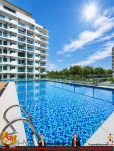 Rooftop rectangular swimming pool with blue tiled water beside a white modern apartment building on a sunny day with a clear blue sky and glass railing.