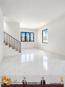 Bright empty living room with white walls, black railing on the staircase to the left, and large windows showing a blue sky and neighboring buildings.