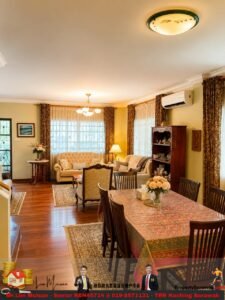 Traditional living and dining room with a patterned tablecloth-covered dining table, six chairs, beige sofa set, and large windows with dark drapes.