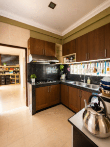 Modern L-shaped kitchen with dark wood cabinets, black granite countertops, and a double sink under a window with colorful utensils hanging above. A stainless steel kettle sits on a nearby table; a doorway leads to a pantry area filled with storage bins.