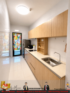 Modern galley kitchen with light wood cabinets, beige countertop, and stainless steel sink along one wall; door with colorful patterned curtain at far end.