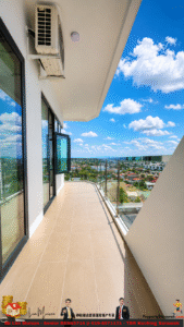 Balcony with tiled floor and glass railing, open doors to a high-rise apartment, overlooking a suburban city under a bright blue sky with clouds. A wall-mounted air conditioner is visible.