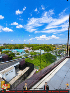 Balcony with glass railing overlooking a green cityscape under a bright blue sky with white clouds.