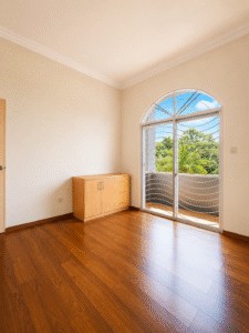 Empty room with wood flooring and a light wood sideboard, large arched window revealing a balcony and green trees outside.
