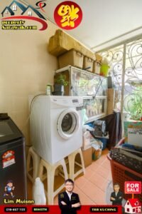 Cramped laundry/storage area with a front-loading washing machine on plastic stools, stacked boxes, and shelves overhead.