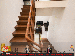 Wooden staircase with a curved handrail descends to a small lounge area with black sofas and a coffee table with a plant, against beige walls.