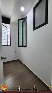 Narrow utility room with white walls and brown floor tiles, featuring a barred window at left and an open black-framed glass door along a white wall. A floor drain is visible near the corner.
