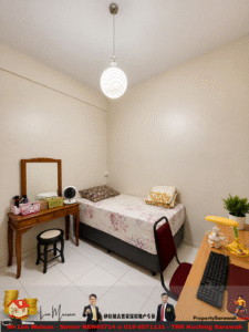 Cozy dorm-style bedroom with a single bed, floral bedsheet, and pillows against beige walls; a wooden vanity with mirror, cosmetics, and a small fan sits at the left; a black stool is near the vanity. A desk with a computer, yellow desk lamp, and a small plant occupies the right side; the room is tiled and softly lit by a pendant ceiling light.