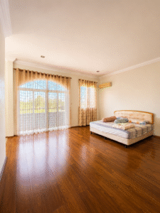 Spacious bedroom with a large arched window covered by sheer beige curtains and a hardwood floor; a low cushioned bed sits against the right wall with blankets and pillows.