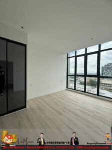 Empty bright room with light wood floors and large black-framed windows showing a city view, white walls, and a glass-panel door on the left.