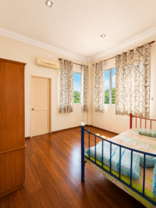 Bright bedroom with a blue metal bed frame, wooden wardrobe, and patterned curtains; warm wood floor and sunlight from two windows.