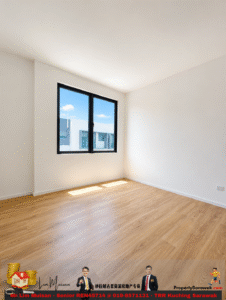 Sunlit, empty room with white walls, light wood floor, and a large black-framed window showing a blue sky outside.