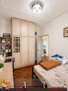 Bedroom with large light wood wardrobe, desk, and bookshelf along left wall; bed with patterned sheets and folded blanket on top, open door to adjoining bathroom.