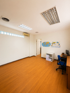 Empty classroom with a wooden floor, desk and computer in the corner, and world maps on the wall near an open door.