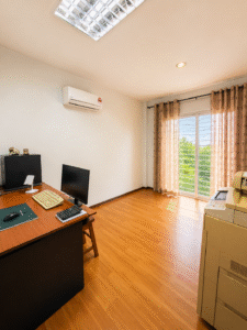 Home office with wooden desk, monitor, keyboard, mouse, and a printer on the right; sunlight through beige curtains.