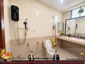 Modern bathroom with beige wall tiles, wall-mounted water heater, handheld shower, toilet, cleaning tools (mop and broom), and long vanity with sink and mirror.