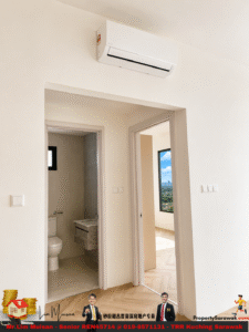 White-walled interior with a wall-mounted air conditioner above a doorway; view into a bathroom with toilet and sink, and an adjacent room with a large window showing a cityscape.