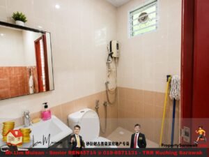 Small beige-tiled bathroom with a white sink, pink soap bottle, and mirror on the left; toilet next to a wall-mounted handheld shower and a window above. A mop and broom stand in the corner, and a real estate watermark runs along the bottom edge.