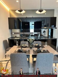 Open-plan kitchen and dining area with a marble table set for four, grey upholstered chairs, black place settings, wine glasses, and a vase of flowers.